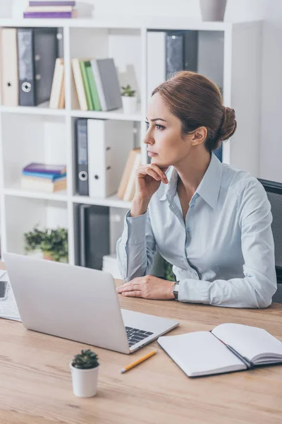 Pensativo hermosa mujer de negocios sentado en el lugar de trabajo y mirando hacia otro lado - foto de stock