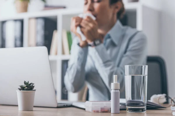 Medikamente stehen auf dem Arbeitstisch, im Hintergrund sitzt eine niesende Geschäftsfrau — Stockfoto