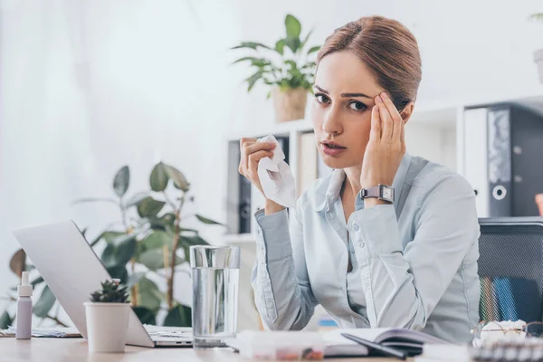 Sick adult businesswoman sitting at workplace with glass of water and looking at camera while touching her head — Stock Photo