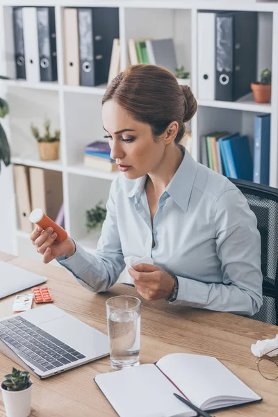 Ill adult businesswoman sitting at workplace with plastic jar of pills and water — Stock Photo