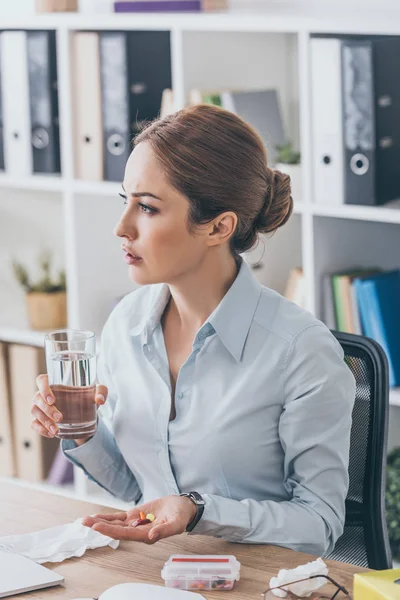 Mujer de negocios enferma pensativa sentada en el lugar de trabajo con pastillas y vaso de agua en la oficina - foto de stock