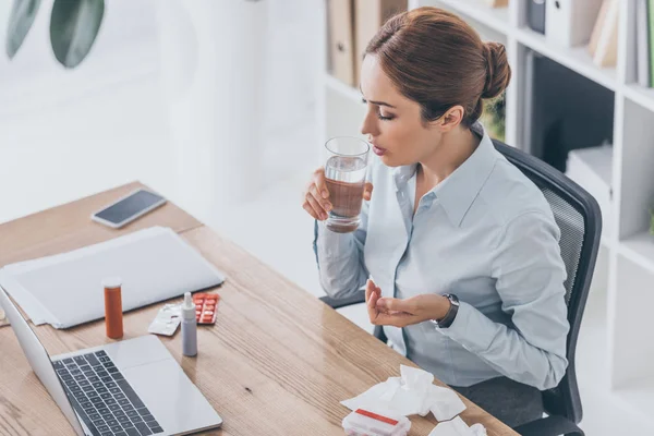 Vista de ángulo alto de la mujer de negocios adulta enferma que toma píldoras en el lugar de trabajo - foto de stock