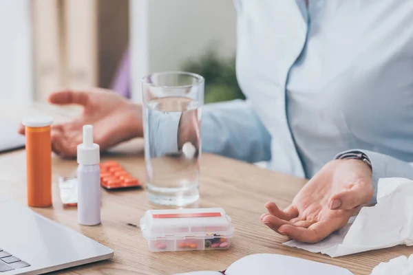 Обрезанный снимок больной предпринимательницы, сидящей на рабочем месте с лекарствами и стаканом воды — стоковое фото