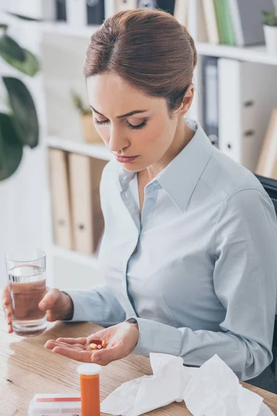 Mulher de negócios doente sentado no local de trabalho com pílulas e copo de água — Fotografia de Stock