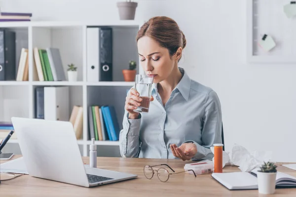 Mujer de negocios adulta enferma sentada en el lugar de trabajo con pastillas y vaso de agua - foto de stock