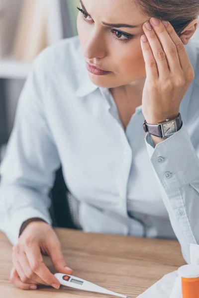 Close-up portrait of diseased businesswoman sitting at workplace and holding electric thermometer — Stock Photo