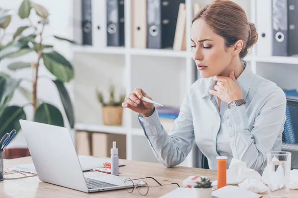 Diseased businesswoman sitting at workplace and holding electric thermometer — Stock Photo
