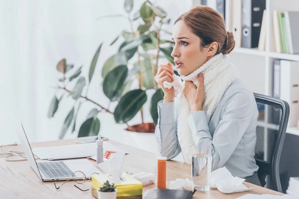 Adult sick businesswoman with flu sitting at workplace in scarf — Stock Photo