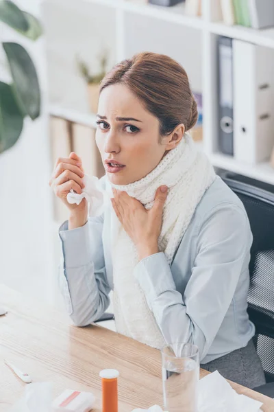 Високий кут зору хворої бізнес-леді в шарфі сидить на робочому місці і дивиться на камеру — стокове фото