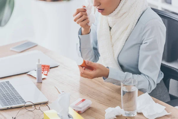 Cropped shot of sick businesswoman in scarf holding container of pills at workplace — Stock Photo