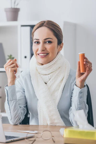 Enferma sonriente empresaria en bufanda sosteniendo contenedor de píldoras en el lugar de trabajo y mirando a la cámara - foto de stock