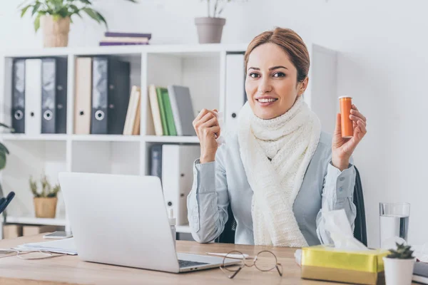 Больная улыбающаяся деловая женщина в шарфе держит контейнер с таблетками в офисе и смотрит в камеру — стоковое фото