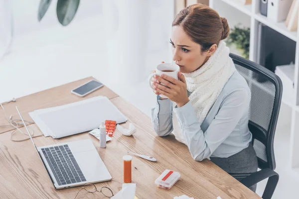 Visão de alto ângulo de empresária doente em cachecol sentado no local de trabalho e beber chá — Fotografia de Stock
