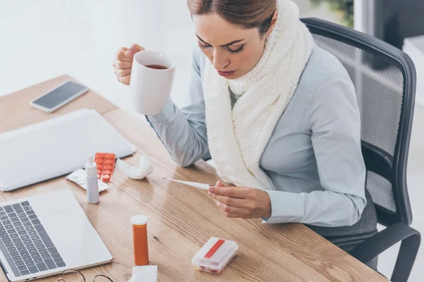 High angle view of sick businesswoman in scarf holding cup of tea and electric thermometer sitting at office — Stock Photo