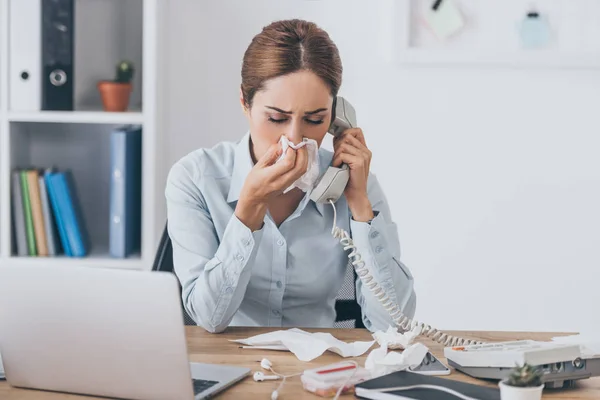 Close-up portrait of ill adult businesswoman with runny nose talking by wired phone at office — Stock Photo
