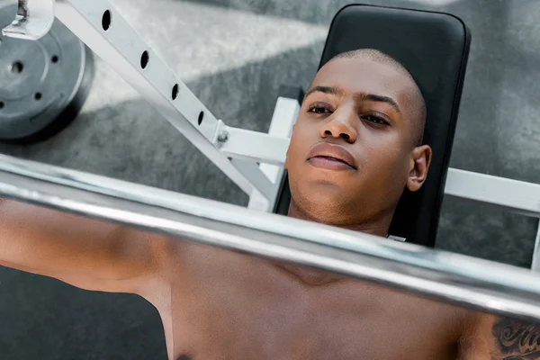 Vue de dessus du jeune sportif afro-américain allongé sur un banc et levant une haltère dans une salle de gym — Photo de stock