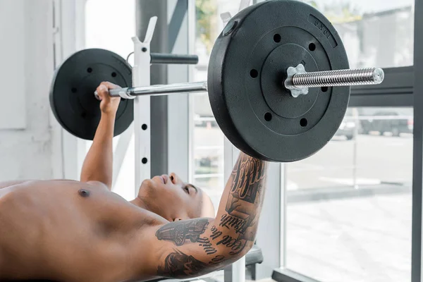 Atlético desnudo en el pecho tatuado deportista acostado en el banco y levantando la barra en el gimnasio - foto de stock