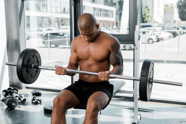 Athletic bare-chested african american sportsman sitting on bench and lifting barbell in gym — Stock Photo