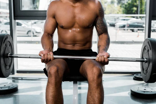 Cropped shot of muscular tattooed sportsman sitting on bench and holding barbell in gym — Stock Photo