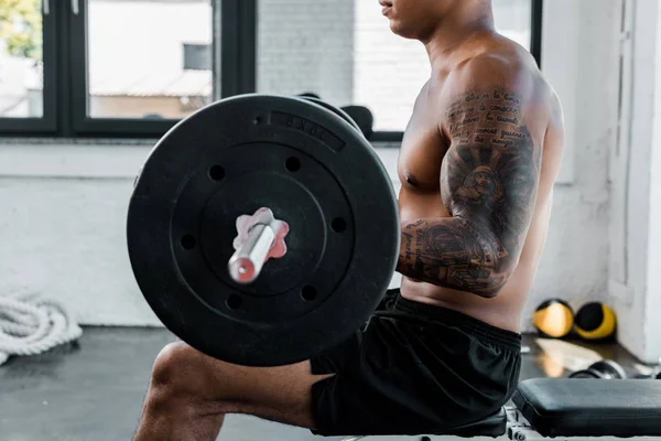 Обрезанный кадр мускулистого спортсмена, сидящего на скамейке и поднимающего штангу в спортзале — стоковое фото
