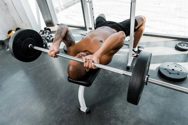 Vista ad alto angolo di muscoloso sportivo afroamericano sdraiato su panchina e sollevamento bilanciere in palestra — Foto stock