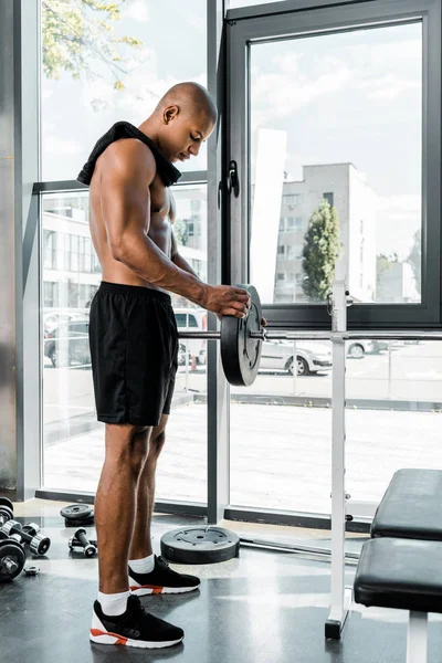 Vue latérale de musclé jeune sportif avec serviette mettre plaque de poids sur haltère dans la salle de gym — Photo de stock