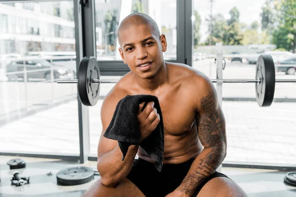 Beau jeune sportif afro-américain musclé tenant une serviette et souriant à la caméra dans la salle de gym — Photo de stock