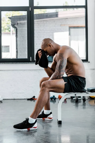 Side view of muscular young sportsman wiping forehead with towel while sitting after workout in gym — Stock Photo