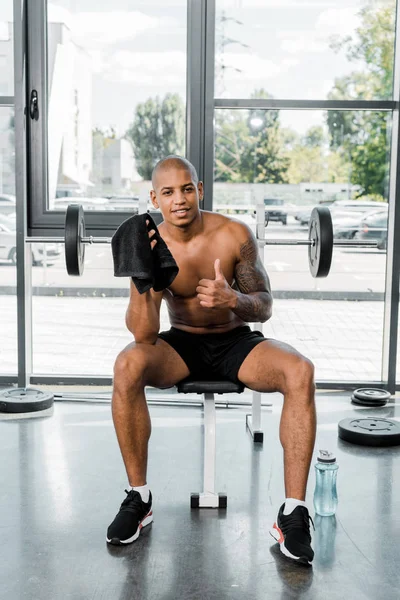 Musculoso joven afroamericano deportista con toalla mostrando el pulgar hacia arriba y sonriendo a la cámara en el gimnasio - foto de stock