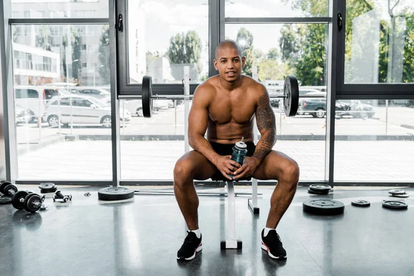 Musculoso joven afroamericano deportista sosteniendo botella de agua y sonriendo a la cámara mientras está sentado en el gimnasio - foto de stock