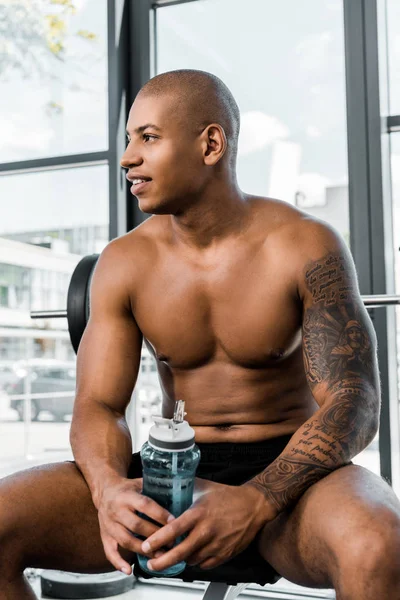 Guapo sonriente joven deportista sosteniendo la botella de agua y mirando hacia otro lado mientras está sentado en el gimnasio - foto de stock