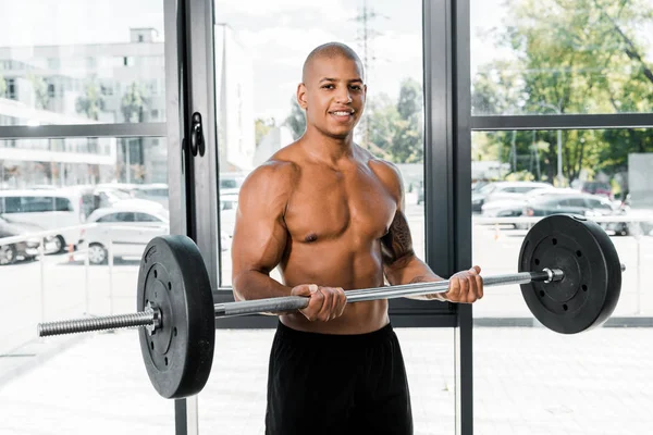Guapo musculoso joven deportista sosteniendo la barra y sonriendo a la cámara en el gimnasio - foto de stock