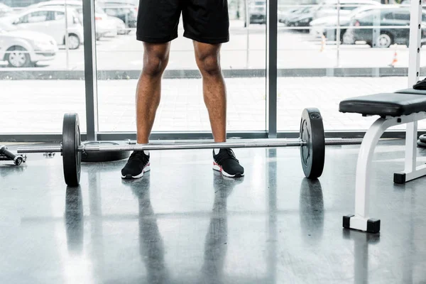 Low section of young sportsman standing near barbell in gym — Stock Photo