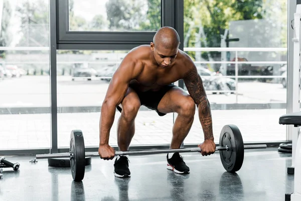 Muscular bare-chested young african american man lifting barbell in gym — Stock Photo