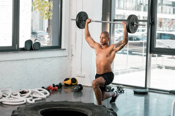 Musculoso desnudo pecho joven deportista levantando la barra y mirando hacia otro lado en el gimnasio - foto de stock