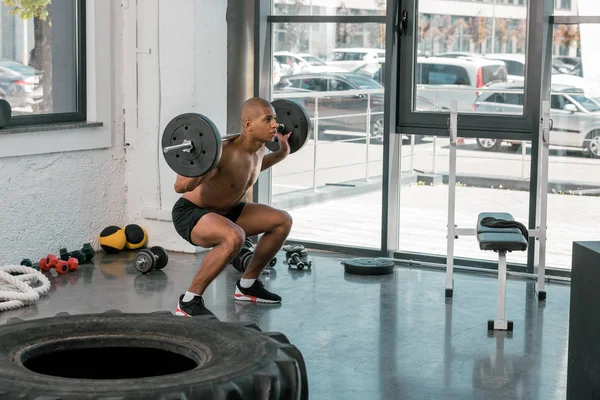 Joven muscular afroamericano hombre levantando la barra y mirando hacia otro lado en el gimnasio - foto de stock