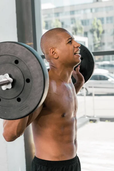 Vista laterale del muscoloso sportivo afroamericano a petto nudo che solleva il bilanciere in palestra — Foto stock