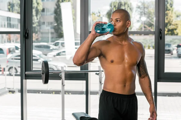 Muskulöser Sportler mit nacktem Oberkörper, der Wasser trinkt und im Fitnessstudio wegschaut — Stockfoto