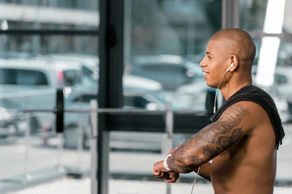 Vista lateral de sonriente deportista sin camisa en auriculares usando smartwatch en el gimnasio - foto de stock