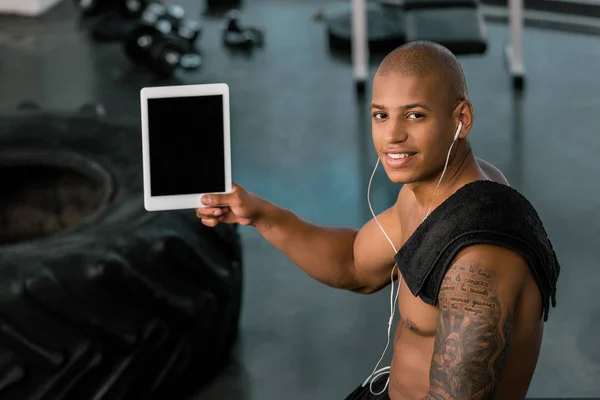 Muscular young sportsman in earphones showing digital tablet with blank screen and smiling at camera in gym — Stock Photo