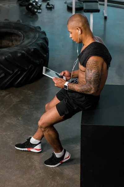 Vue grand angle du jeune sportif afro-américain dans les écouteurs en utilisant une tablette numérique dans la salle de gym — Photo de stock