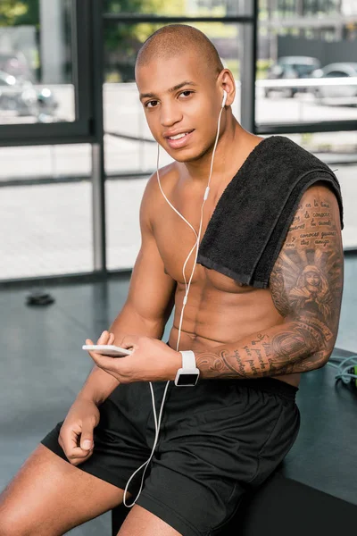 Muscular young sportsman in earphones using smartphone and smiling at camera in gym — Stock Photo