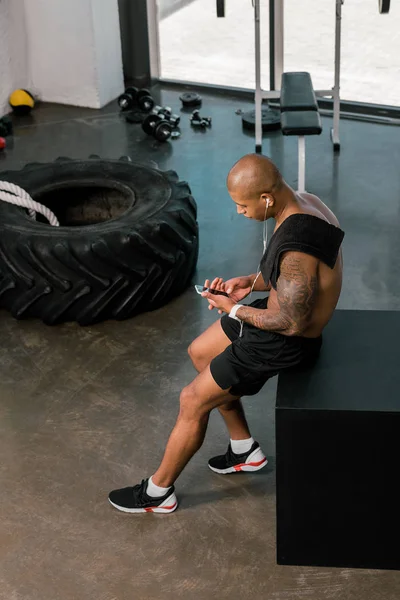 Высокий угол обзора мускулистой молодой спортсмен в наушниках сидя и используя смартфон в тренажерном зале — стоковое фото