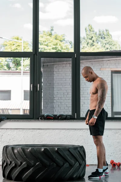 Seitenansicht eines muskulösen jungen afrikanisch-amerikanischen Mannes, der im Fitnessstudio steht und auf Reifen schaut — Stockfoto