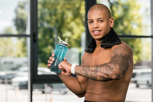 Sonriente joven musculoso hombre sosteniendo botella de agua y comprobando smartwacth en gimnasio - foto de stock