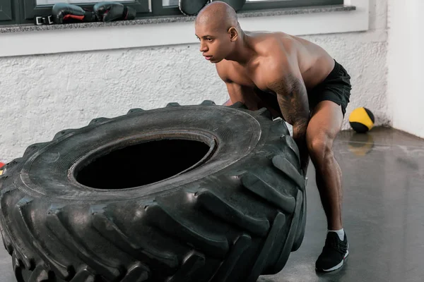 Young muscular bare-chested african american sportsman training with tyre in gym — Stock Photo