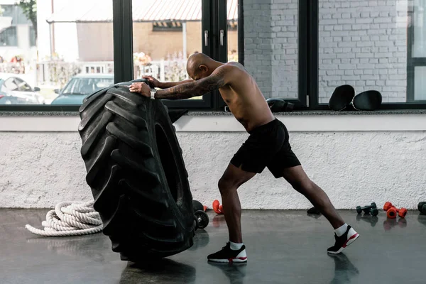 Vue latérale d'entraînement sportif afro-américain torse nu musclé avec pneu dans la salle de gym — Photo de stock