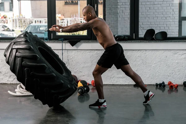 Vista lateral del hombre musculoso sin camisa haciendo ejercicio con neumático en el gimnasio - foto de stock