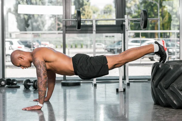 Vista lateral del hombre americano africano muscular haciendo flexiones con neumático en el gimnasio - foto de stock