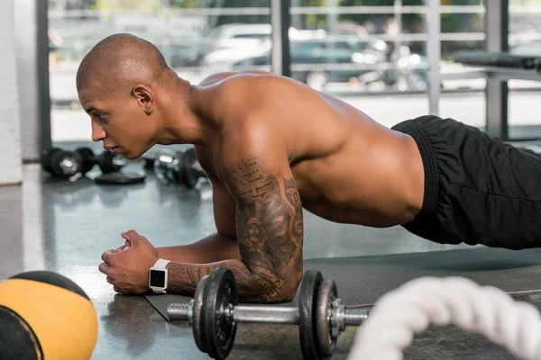 Vista lateral del deportista afroamericano tatuado haciendo tablón en el gimnasio - foto de stock